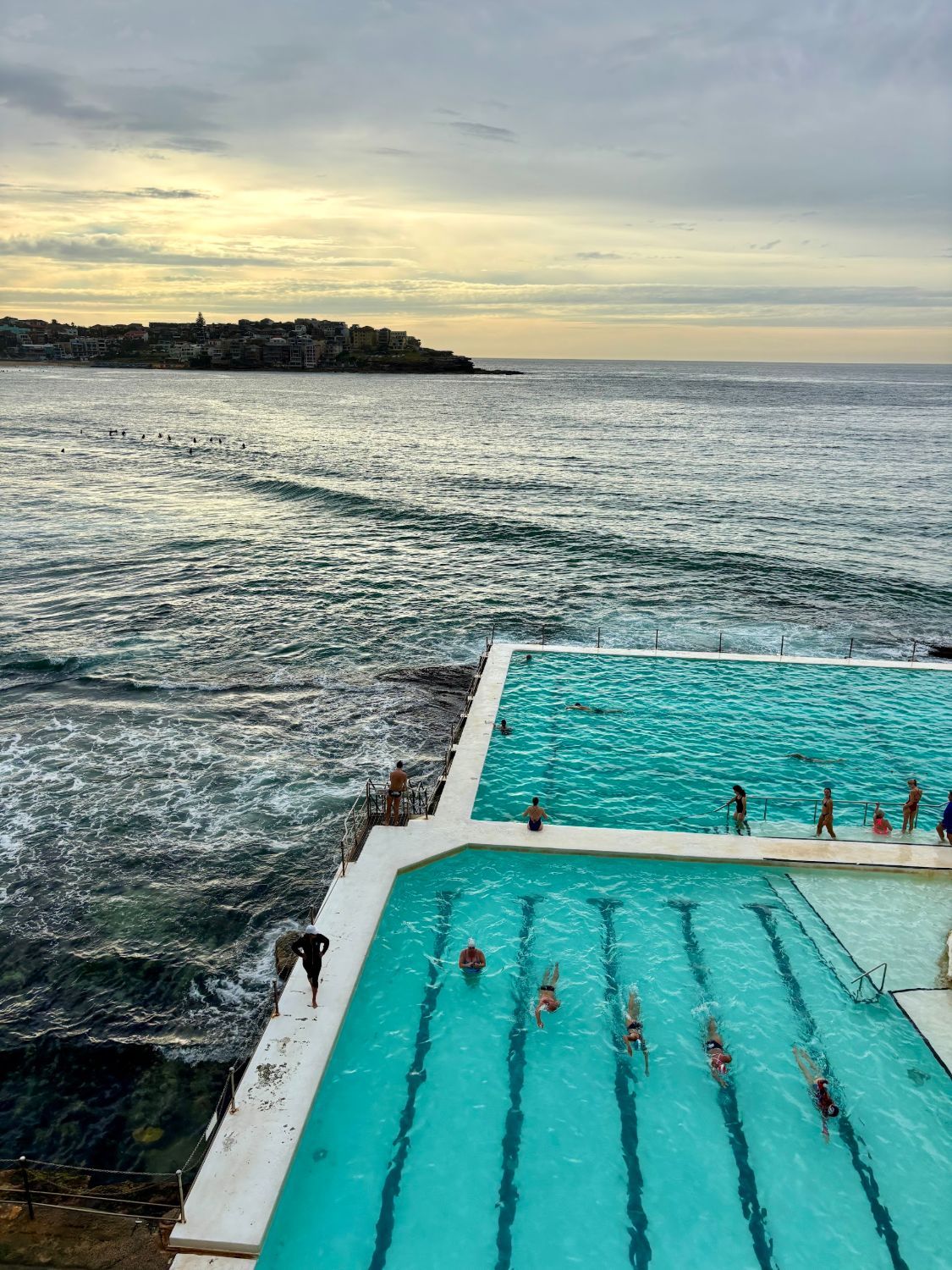 Swimmers doing laps in the two Bondi Icebergs pools beside the sea, with Bondi Beach and the North Bondi headland behind under a low, cloudy sky